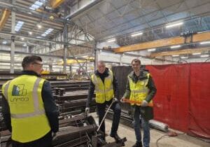 Trabalhadores vestindo coletes de segurança estão em uma oficina industrial cercada por materiais metálicos.